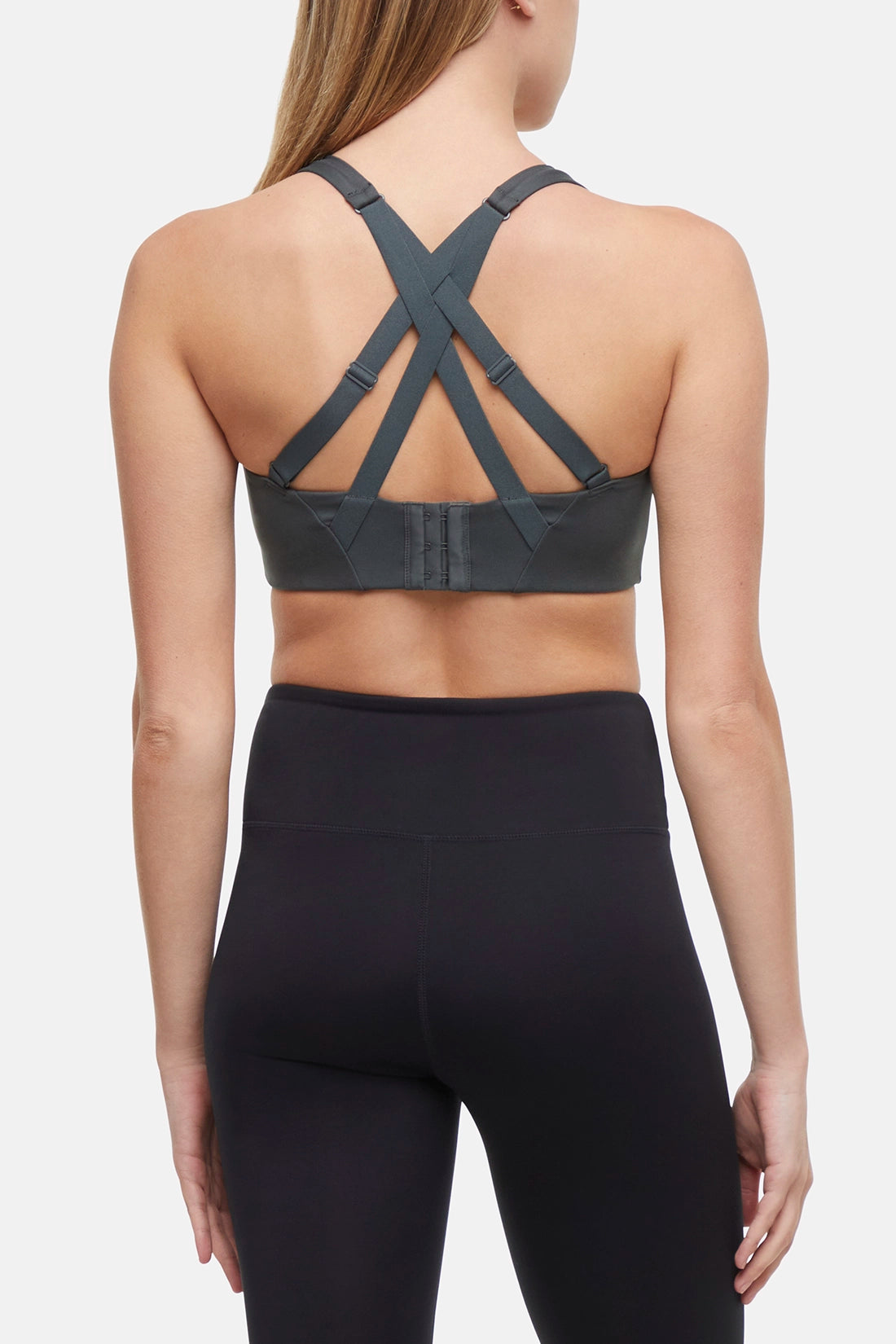 Back view of woman wearing a gray sports bra with criss-cross straps and black leggings on a white background.