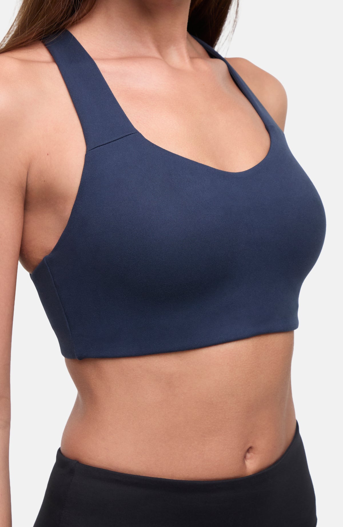 A close-up of a woman wearing a navy blue sports bra with wide straps and a scoop neckline, paired with black bottoms, against a light gray background.