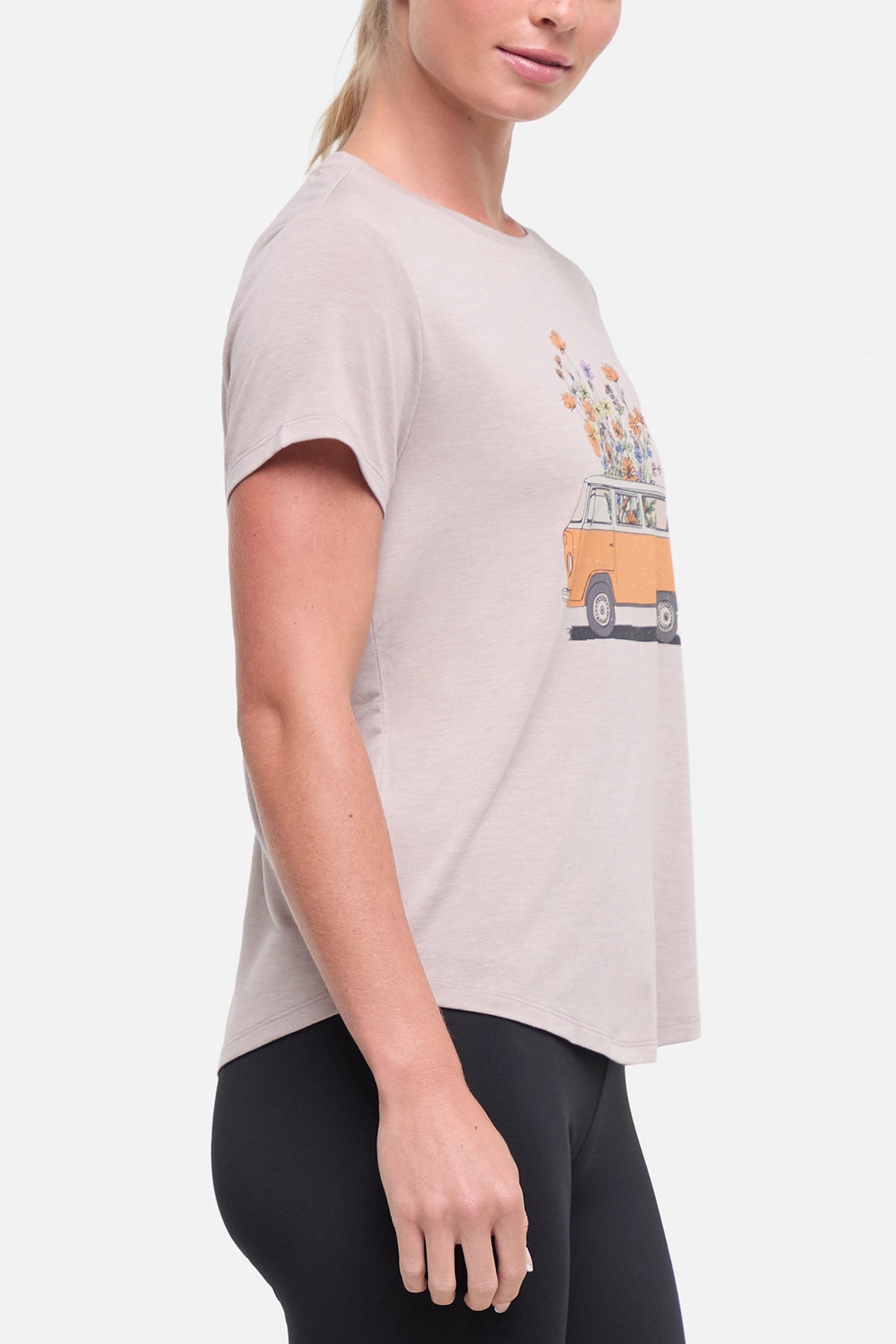 Side view of woman wearing a beige t-shirt with a graphic of an orange van and flowers on a .white background.