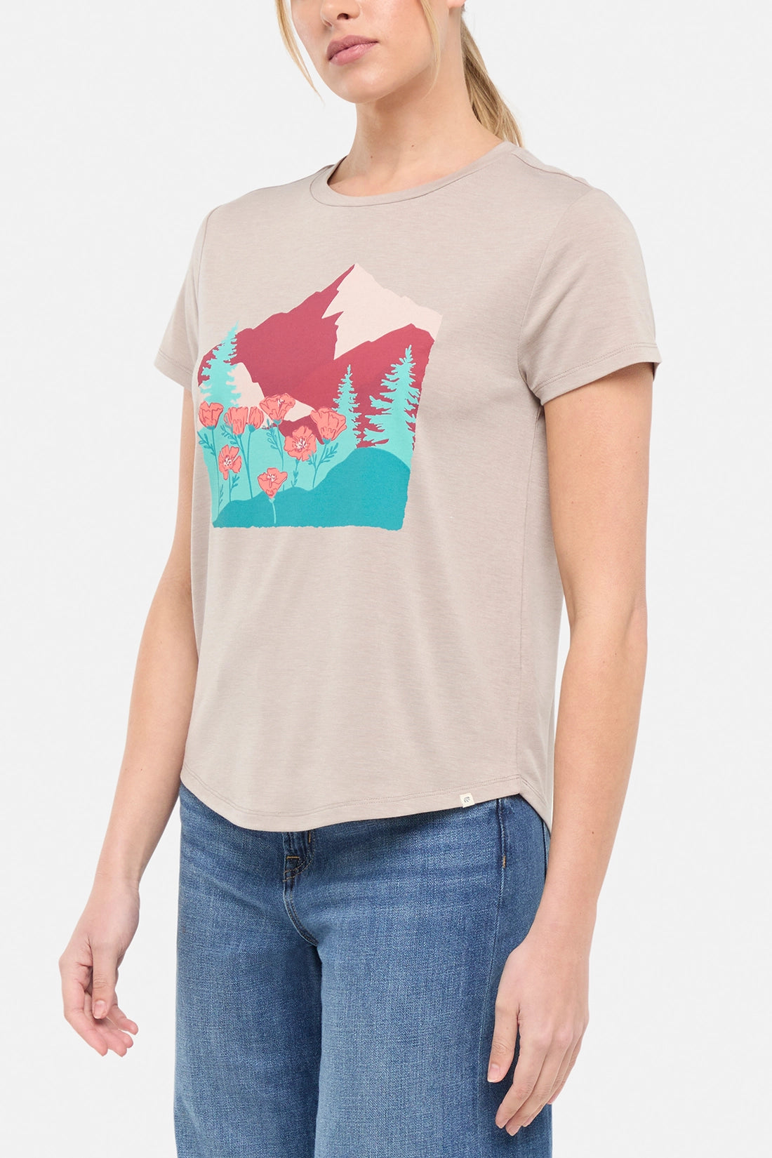 Side view of woman wearing a beige t-shirt with a graphic design of trees mountains, and poppy flowers, paired with blue jeans on white background.