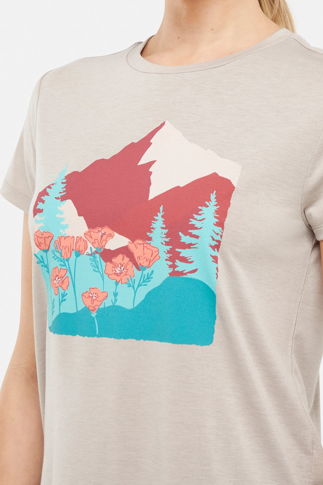 Close up view of woman wearing a beige t-shirt with a graphic design of trees mountains, and poppy flowers, paired with blue jeans on white background.