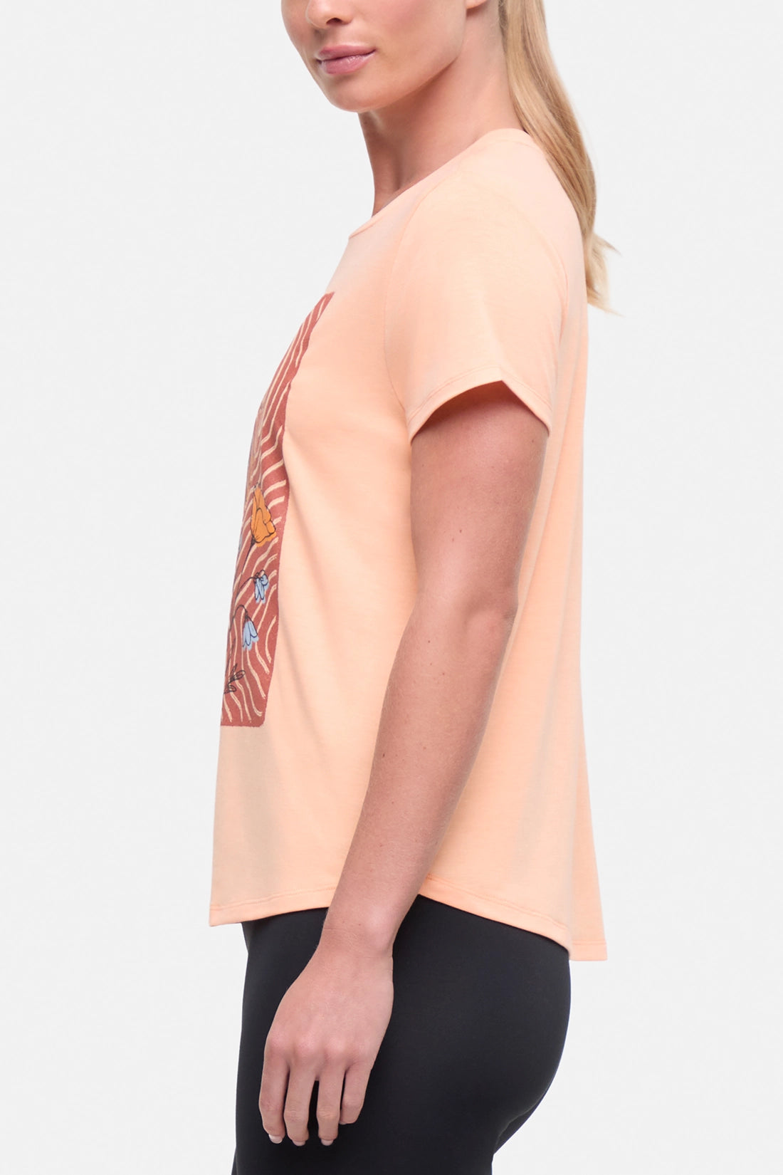 Side view of woman wearing a peach t-shirt with floral graphic on a light gray background.