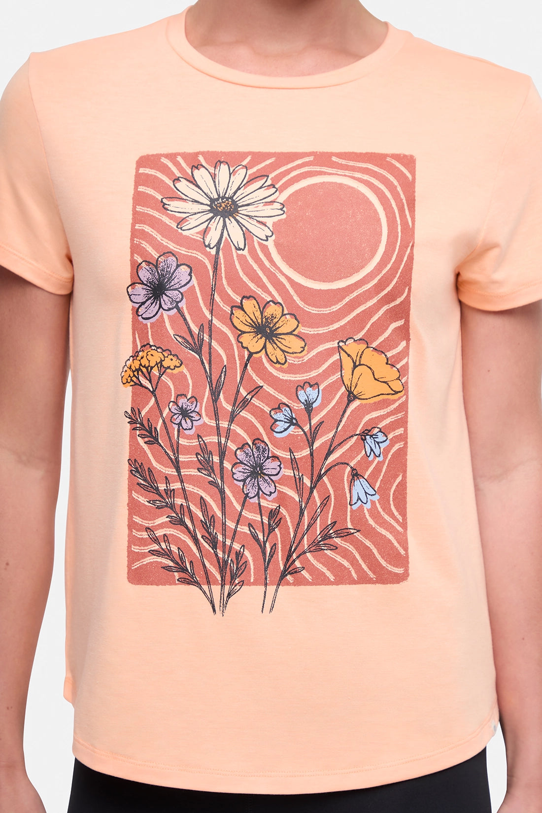 Close up front view of woman wearing a peach t-shirt with floral graphic on a light gray background.