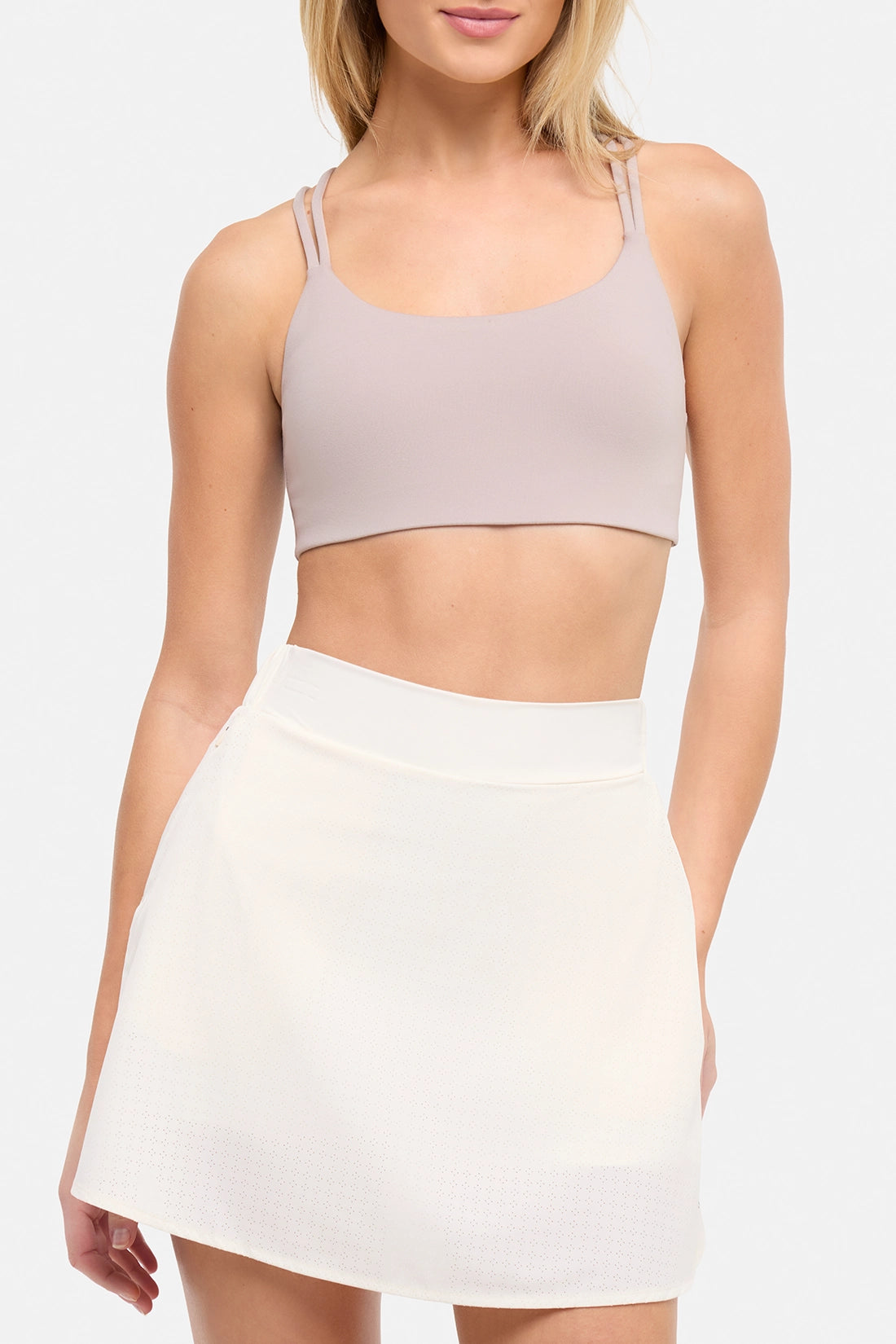 Front view of woman wearing a light grey-beige sports bra and white skort on a white background.