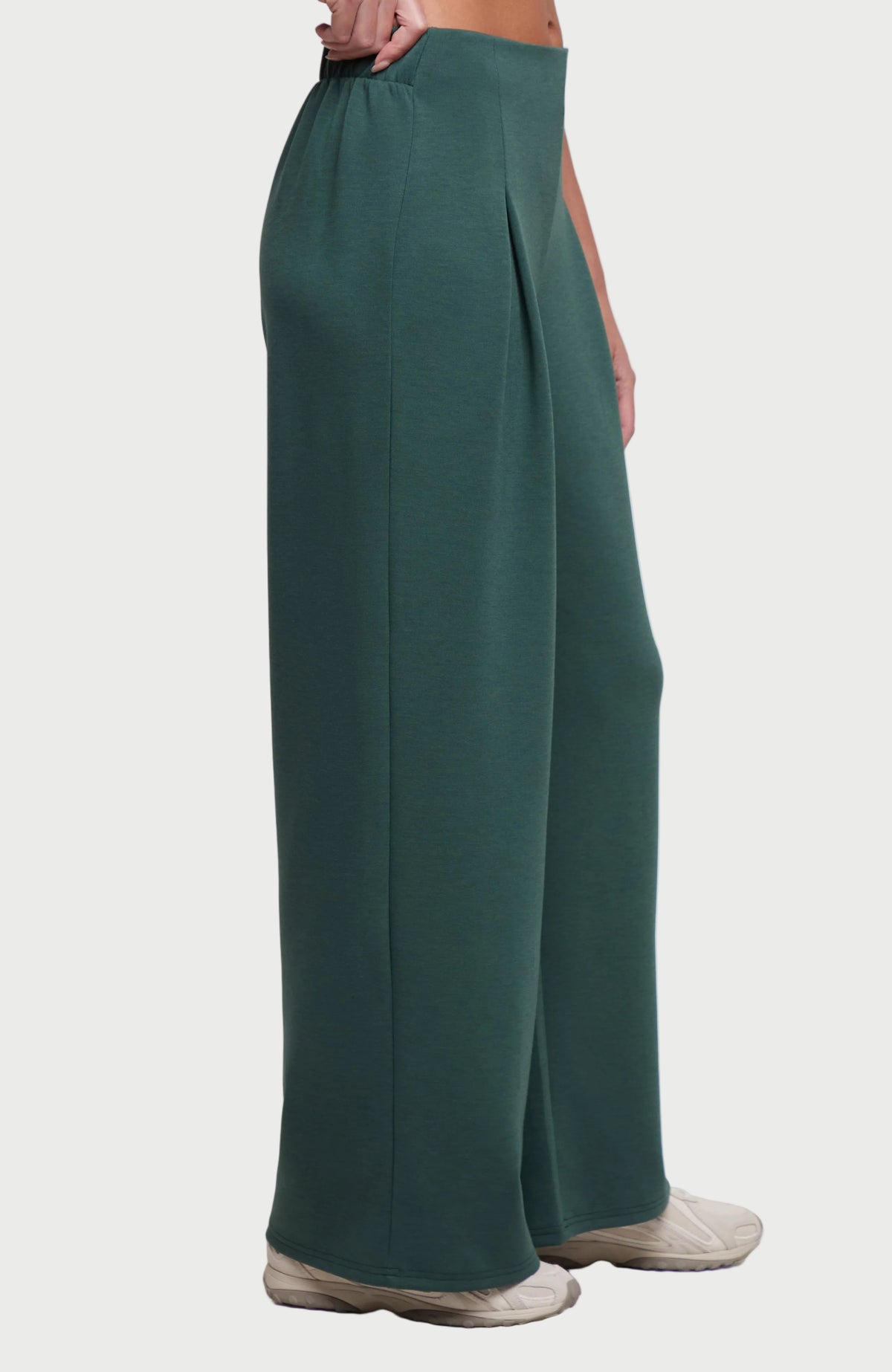 A side view of a person wearing dark green wide-leg pleated pants with light beige slip-on shoes, shown against a plain white background.