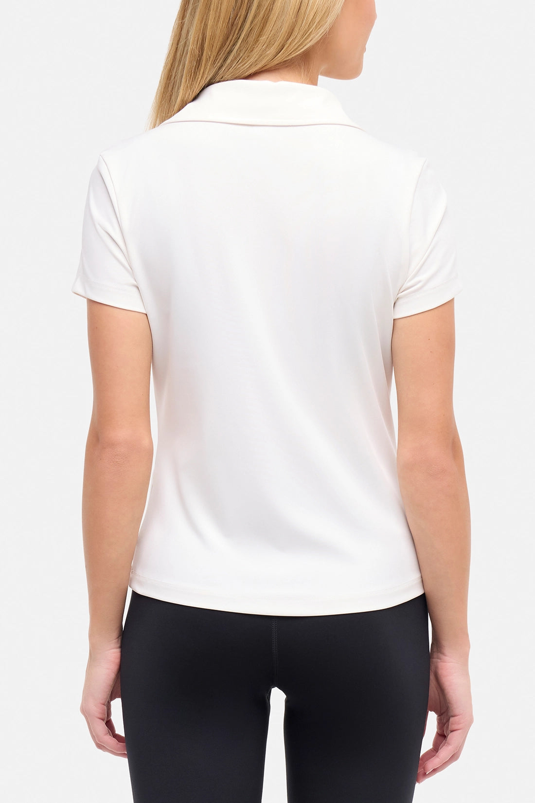 Back view of woman wearing a white short-sleeve polo shirt and black pants on a plain background.