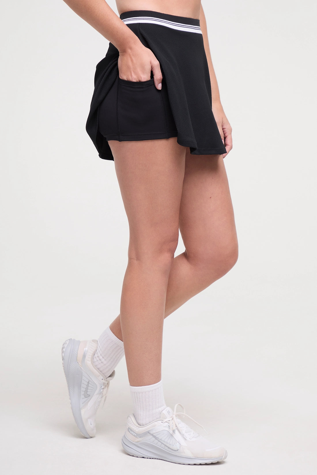 Side view of black tennis skirt with white stripe waistband and inner short pocket worn by a woman on a white background.
