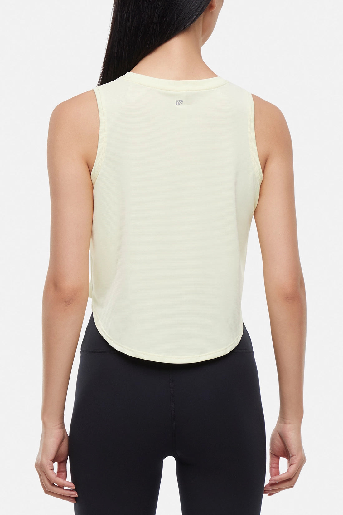 Back view of woman wearing a light yellow sleeveless top and black leggings on a light gray background.