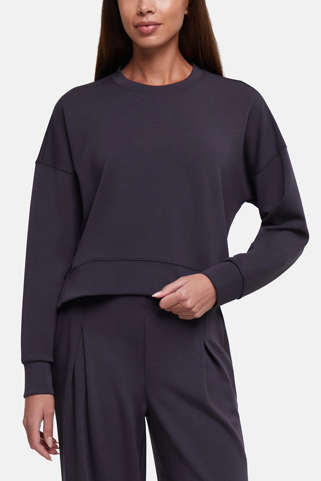 A woman wearing a dark charcoal gray cropped long-sleeve sweatshirt and matching wide-leg sweatpants against a light gray studio background.