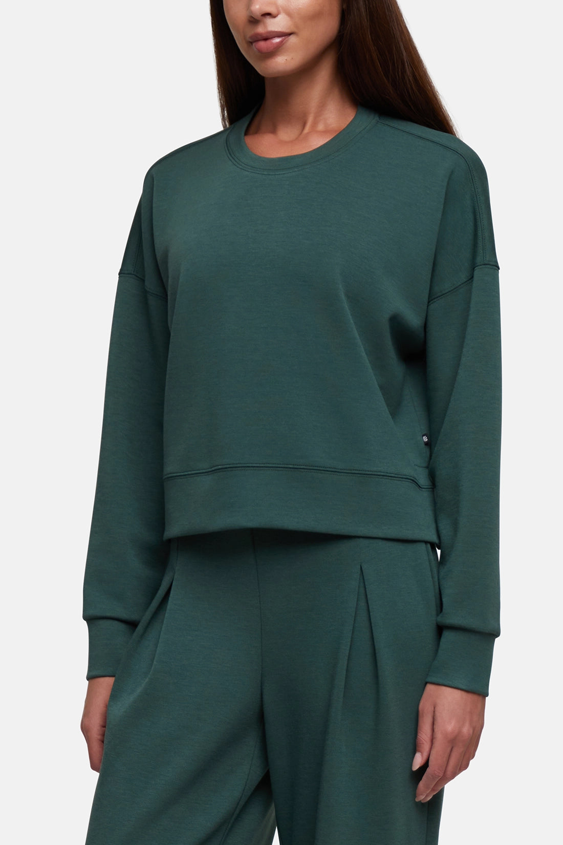 A woman wearing a dark green crewneck sweatshirt and matching wide-leg sweatpants against a light gray studio background.