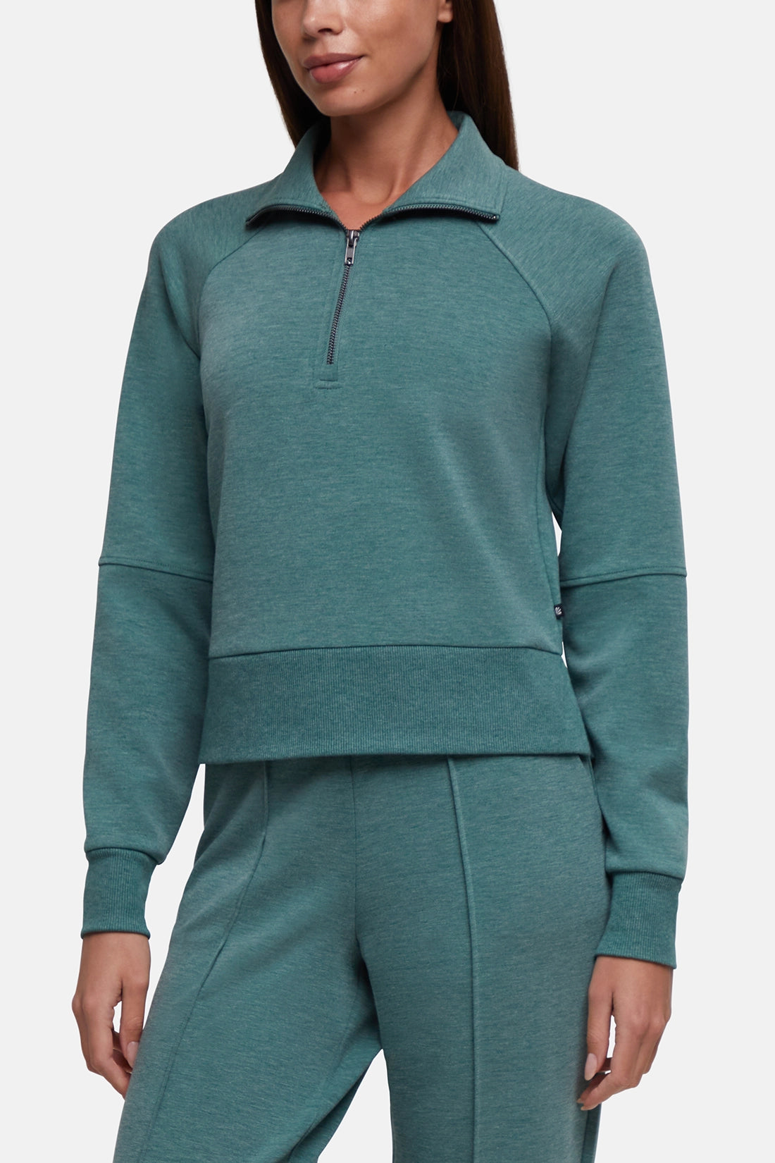 A woman wearing a matching teal green half-zip pullover sweatshirt and jogger pants set, shown from the nose down against a clean white studio background.