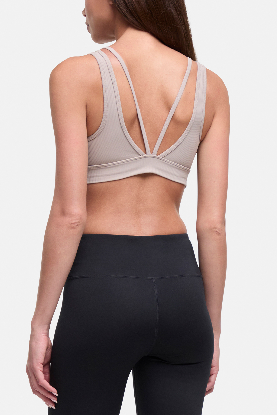 Rear view of a woman wearing a light beige sports bra with a crisscross open-back design and black leggings, against a light gray studio background.