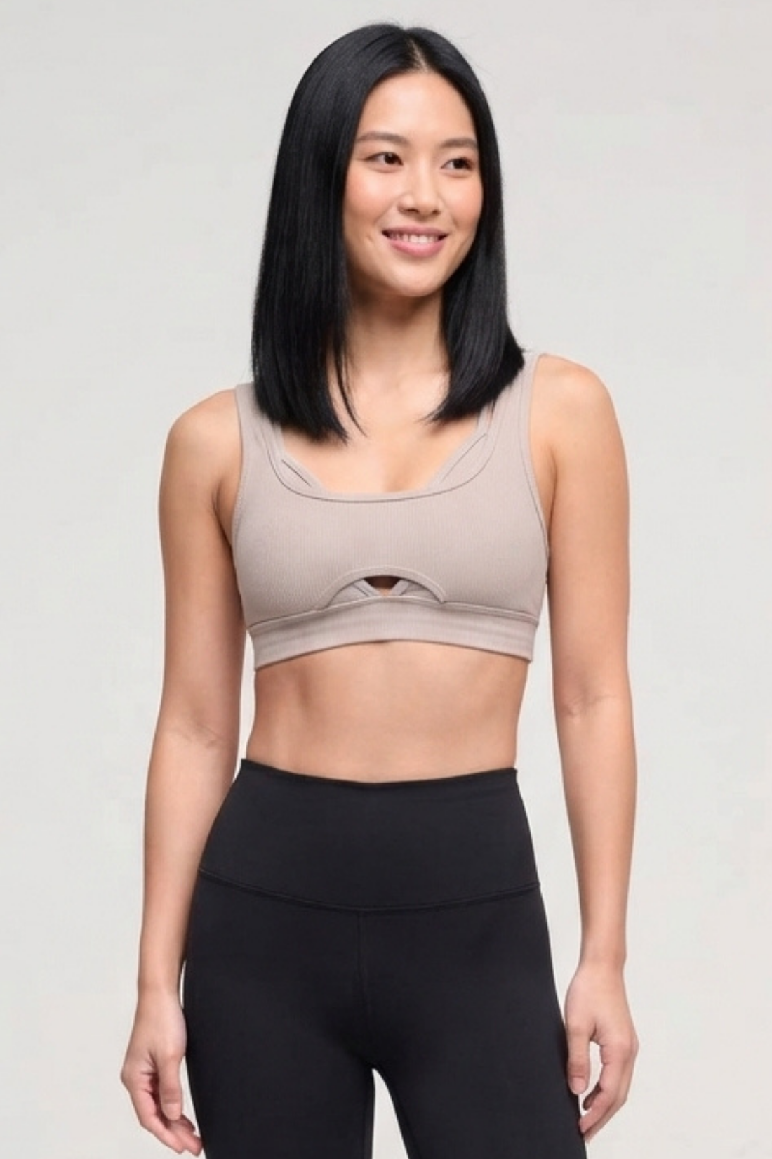 A woman modeling a light taupe-beige athletic sports bra with a small front cutout, paired with black high-waisted leggings, against a light gray studio background.