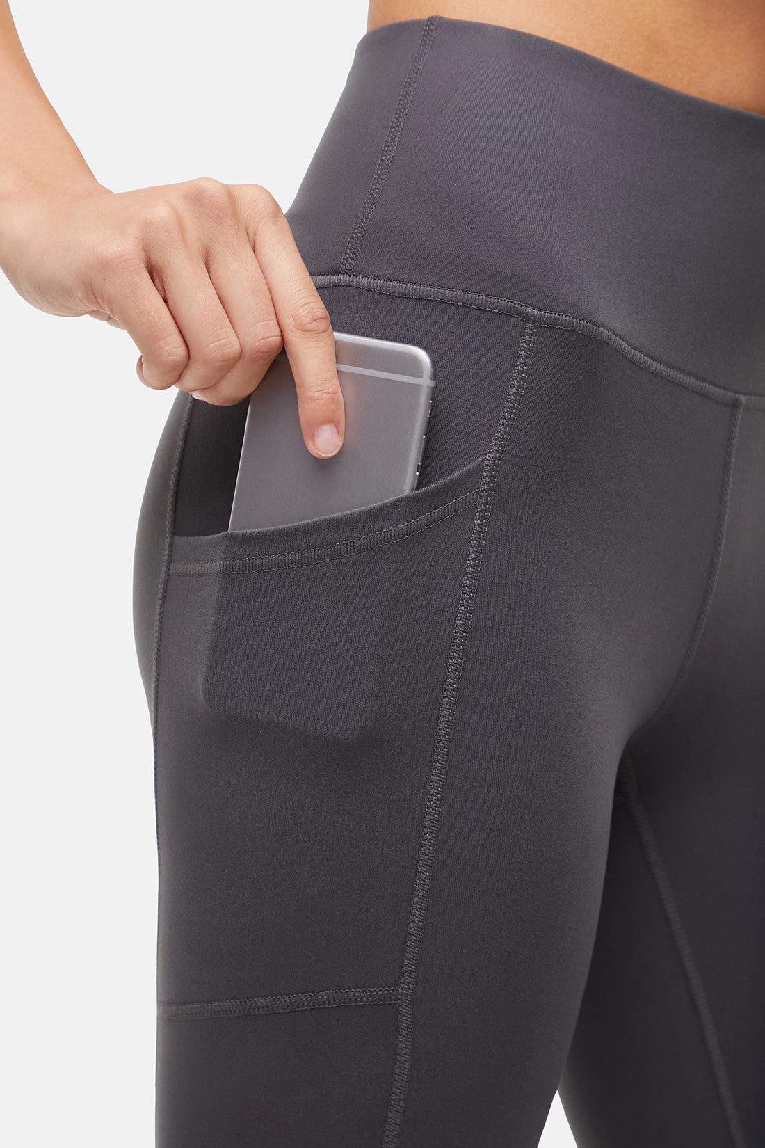 Close up side view of woman wearing dark gray leggings with a side pocket holding a phone on a white background. 