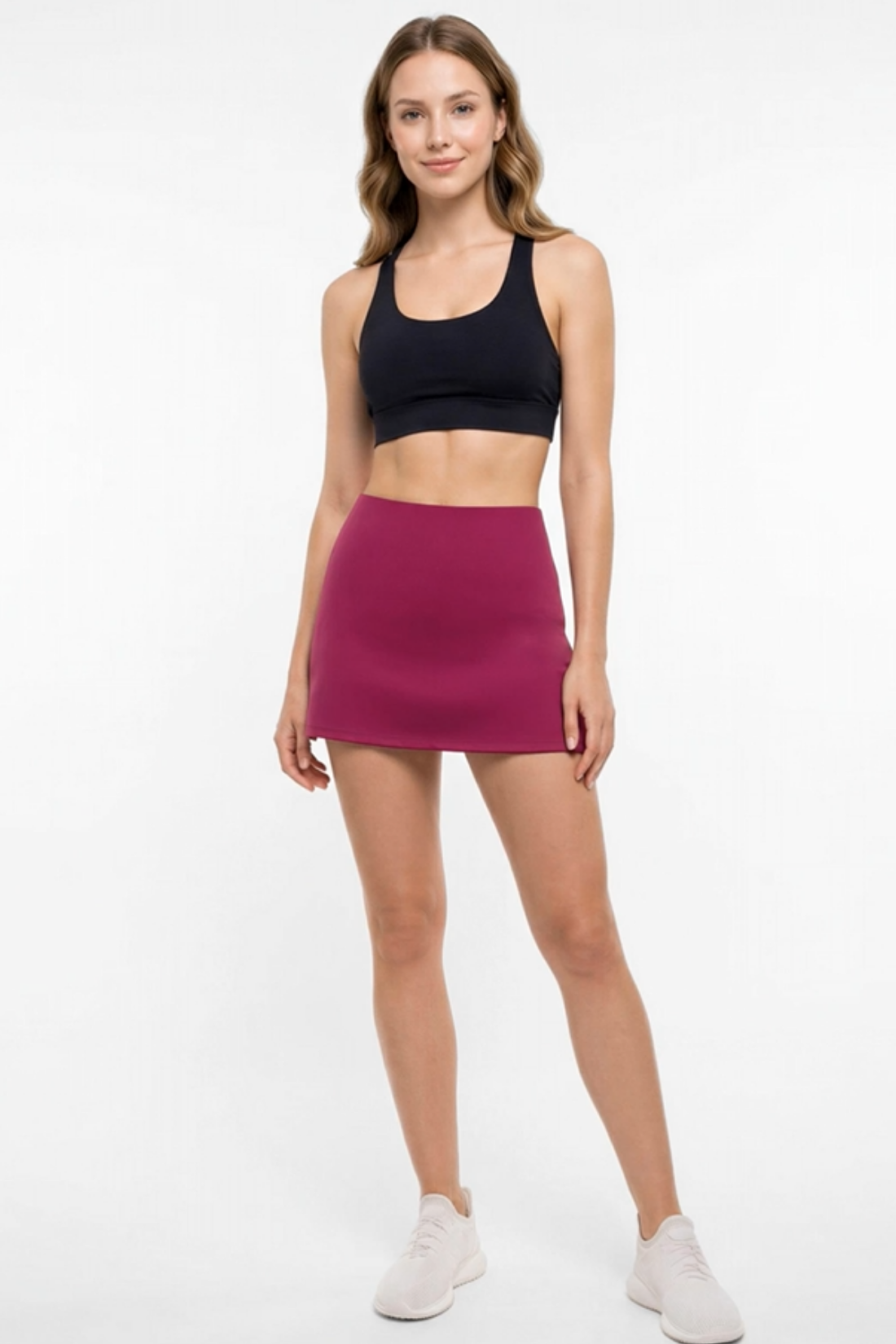 A woman modeling a high-waisted, sleeveless magenta athletic mini skirt paired with a black sports bra and white sneakers, set against a plain white studio background.