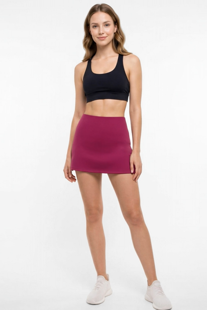 A woman modeling a high-waisted, sleeveless magenta athletic mini skirt paired with a black sports bra and white sneakers, set against a plain white studio background. 