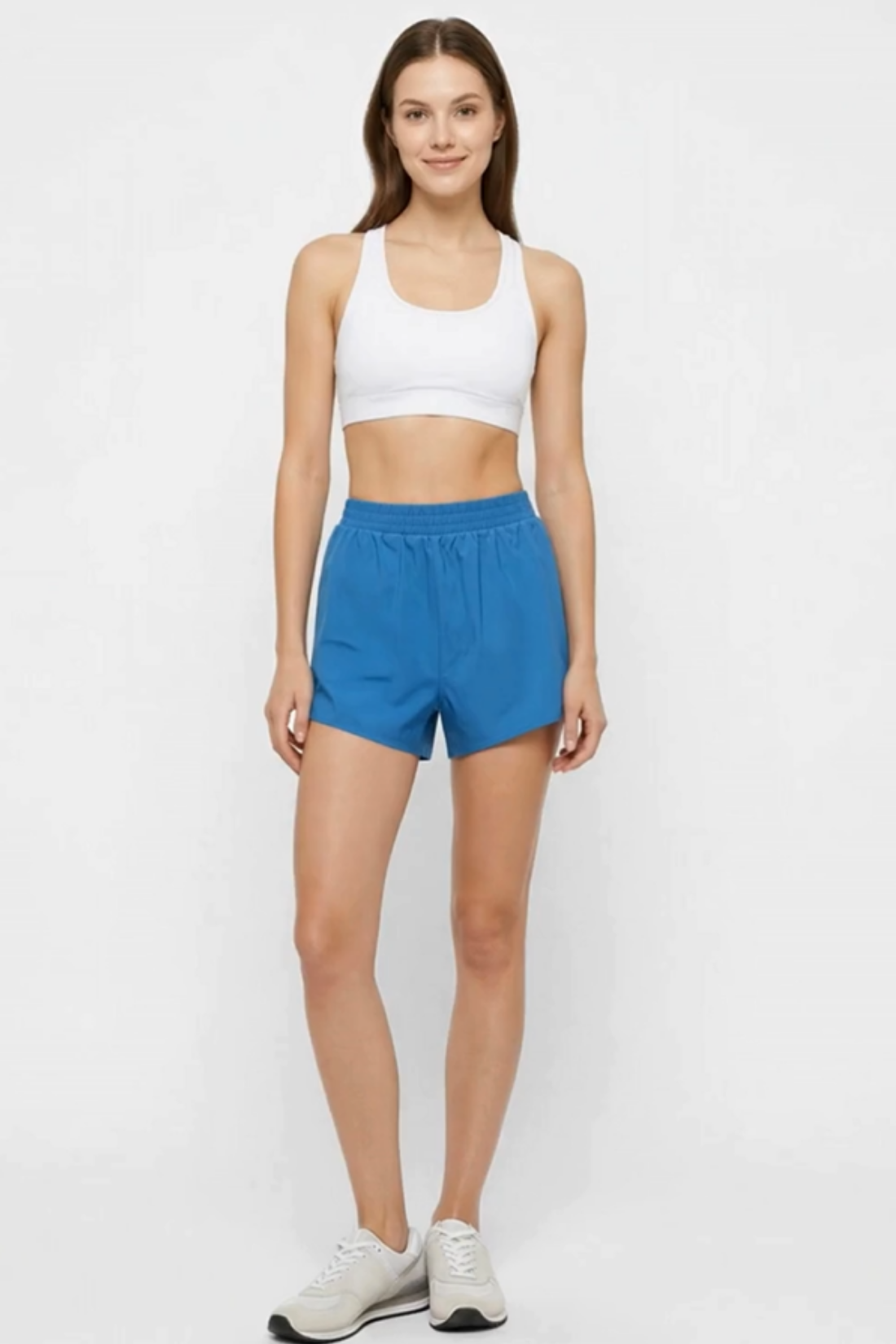 A woman modeling a white sports bra and bright blue athletic shorts against a plain white studio background. 
