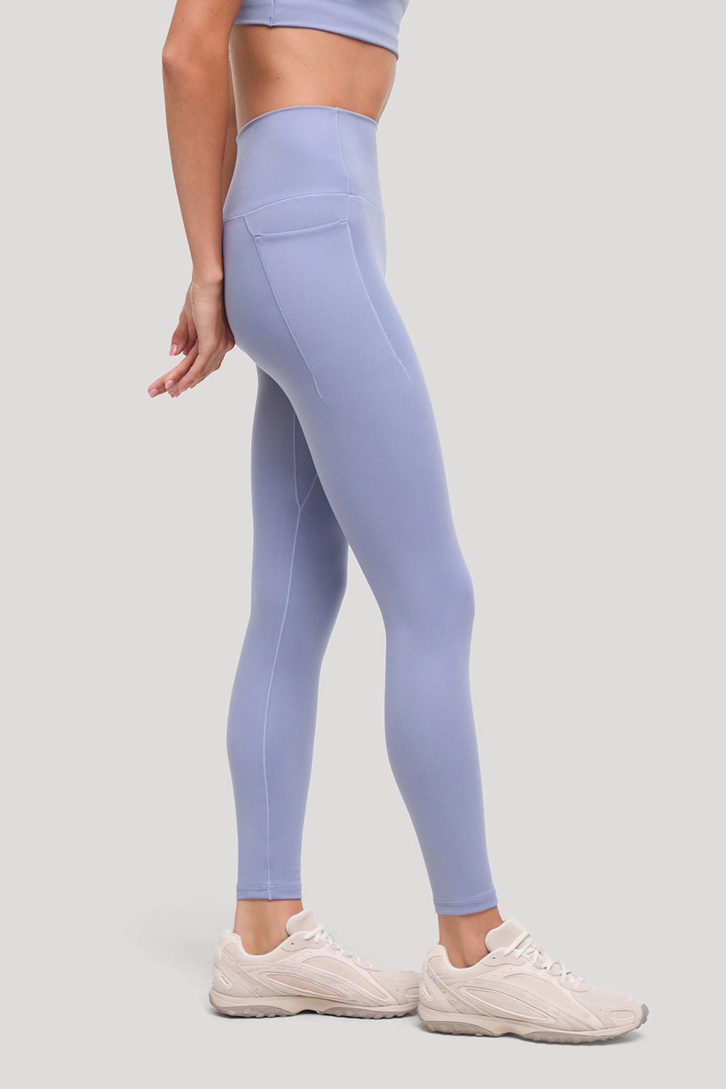 A model wearing light periwinkle blue high-waisted leggings with a side pocket, paired with beige sneakers, shown from a side-back angle against a solid light gray background. #color_tempest