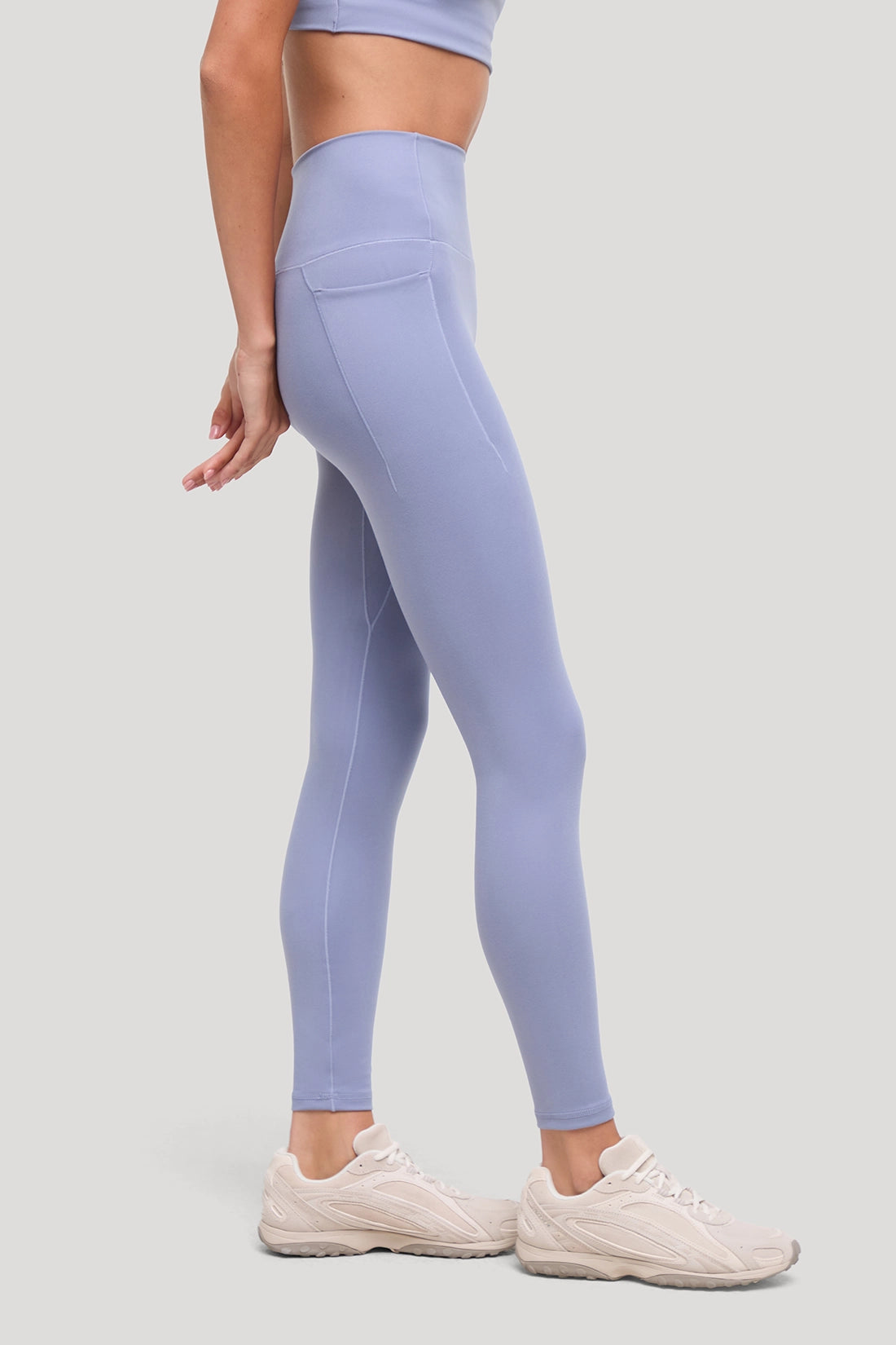 A model wearing light periwinkle blue high-waisted leggings with a side pocket, paired with beige sneakers, shown from a side-back angle against a solid light gray background. #color_tempest