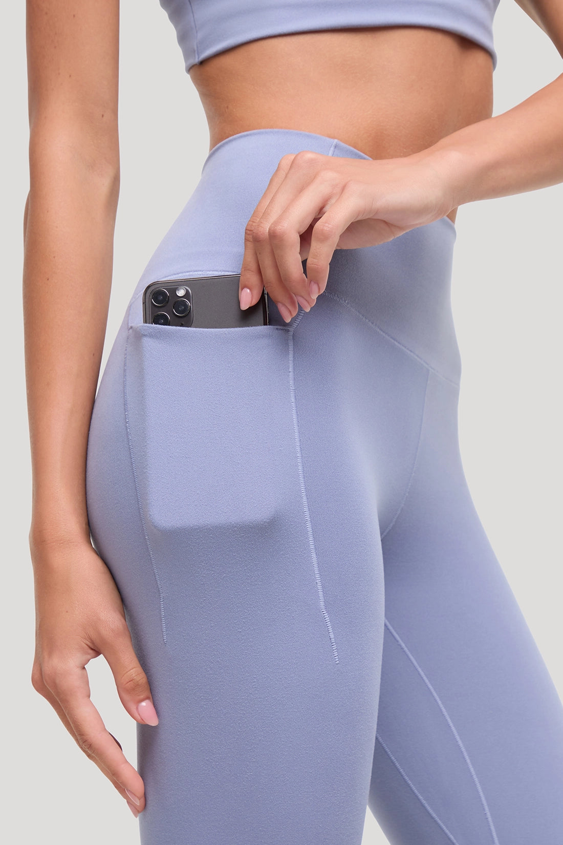 A close-up of a model wearing light periwinkle blue high-waisted leggings with a side pocket containing a smartphone, demonstrating its pocket utility. #color_tempest