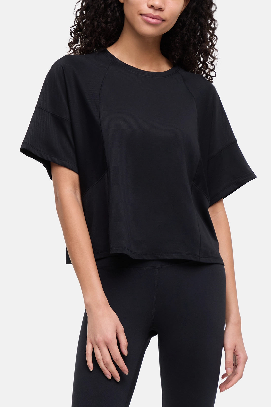 A woman wearing a black short-sleeve crop top with raglan sleeves and black leggings, photographed against a light gray studio background. 
