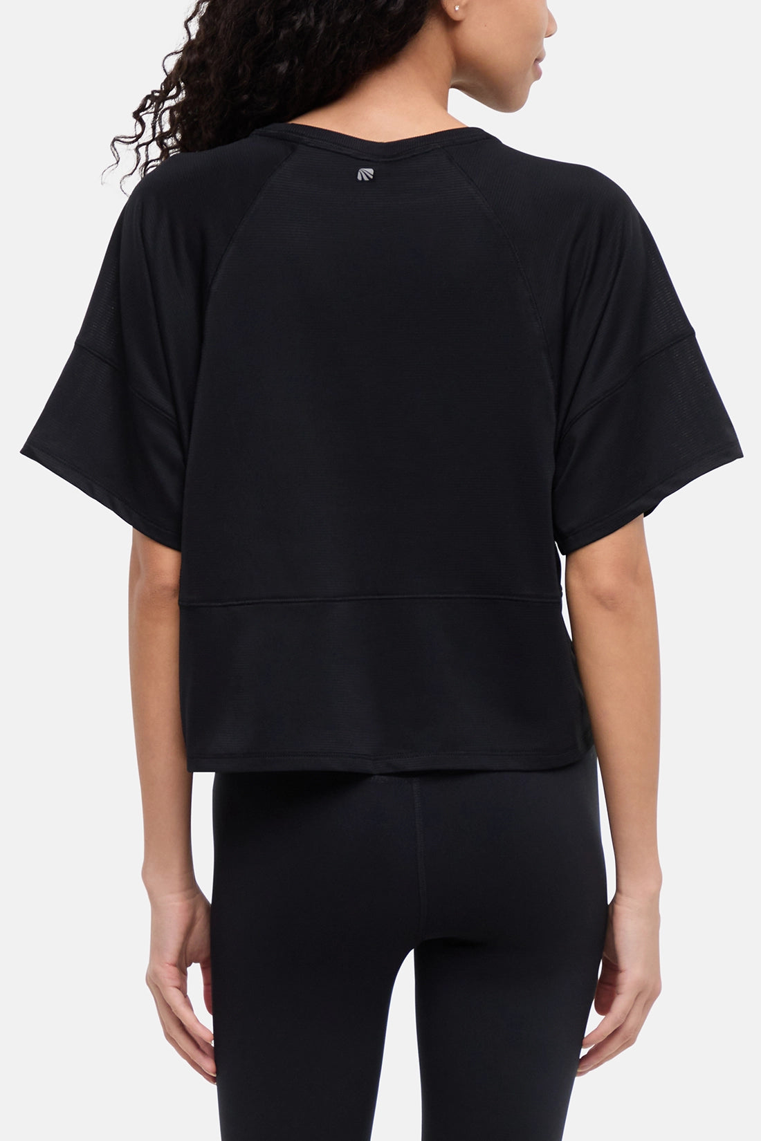 A woman shown from the back wearing a black short-sleeve athletic top and black leggings against a light gray studio background. 