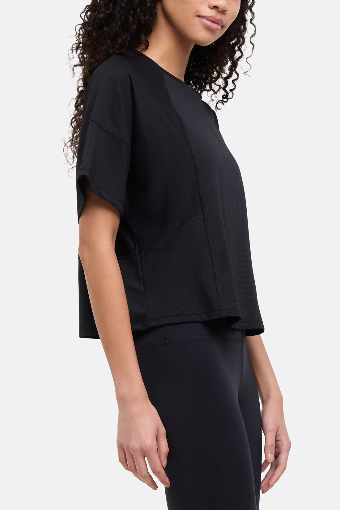 A woman wearing a black short-sleeve crop top with draped, layered sleeves and matching black leggings, shown from the side against a light gray studio background. 