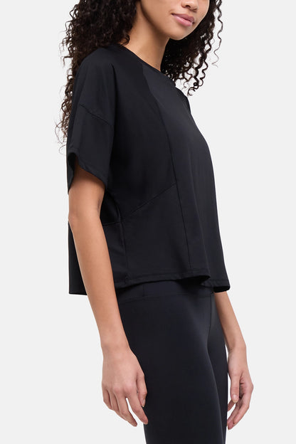 A woman wearing a black short-sleeve crop top with draped, layered sleeves and matching black leggings, shown from the side against a light gray studio background. 