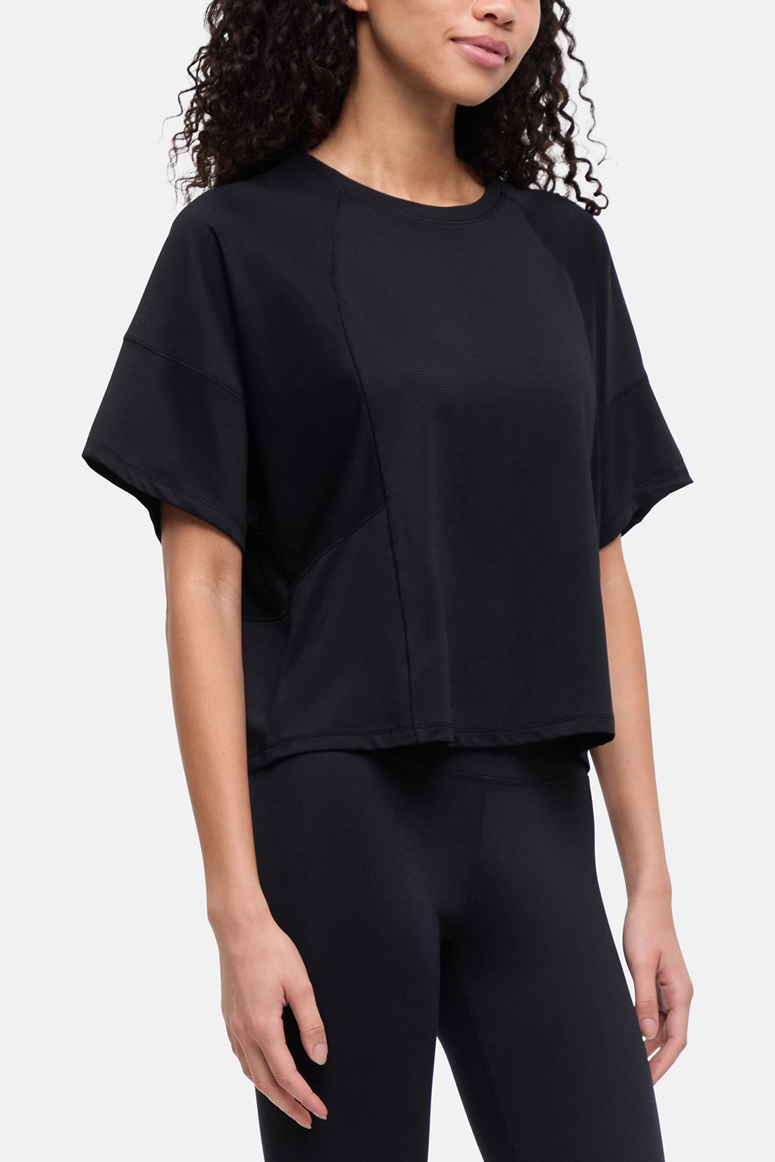A woman wearing a black short-sleeve crop top with a relaxed, draped silhouette and black leggings, photographed against a clean light gray studio background. 