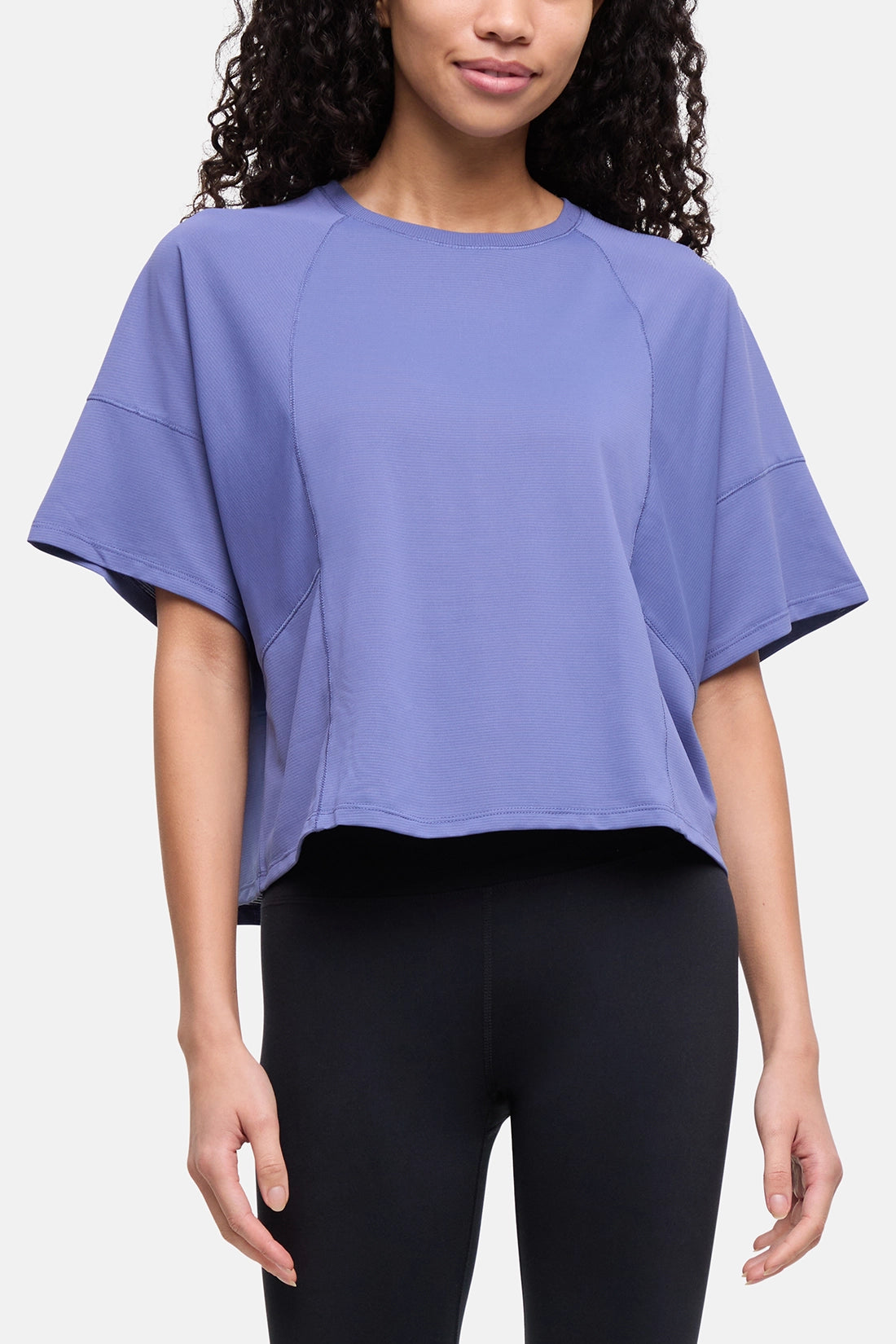 A woman wearing a lavender-purple short-sleeve crop top with raglan sleeves and black leggings, shown from the waist up against a light gray studio background. 