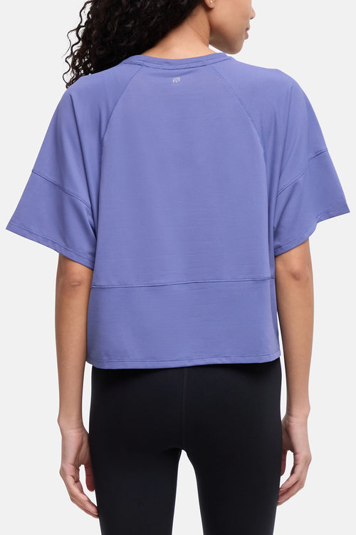 A woman shown from the back wearing a loose lavender-purple athletic top with raglan sleeves and black leggings, against a plain light gray studio background. #color_gray-blue