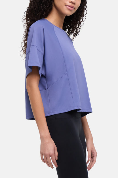 A woman wearing a lavender-purple short-sleeve crop top with diagonal seam details, paired with black leggings, shown against a plain light gray background. 