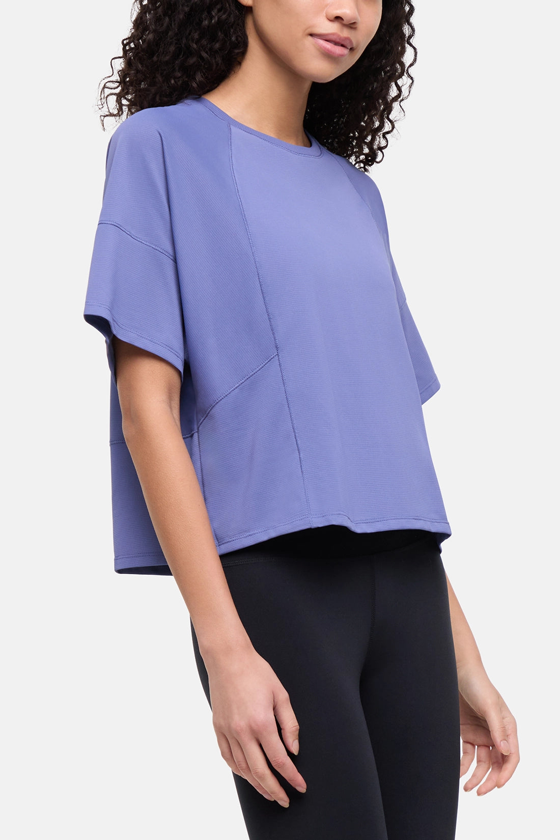 A woman wearing a lavender-purple short-sleeve athletic top with diagonal paneling and black leggings, photographed against a clean light gray studio background. 