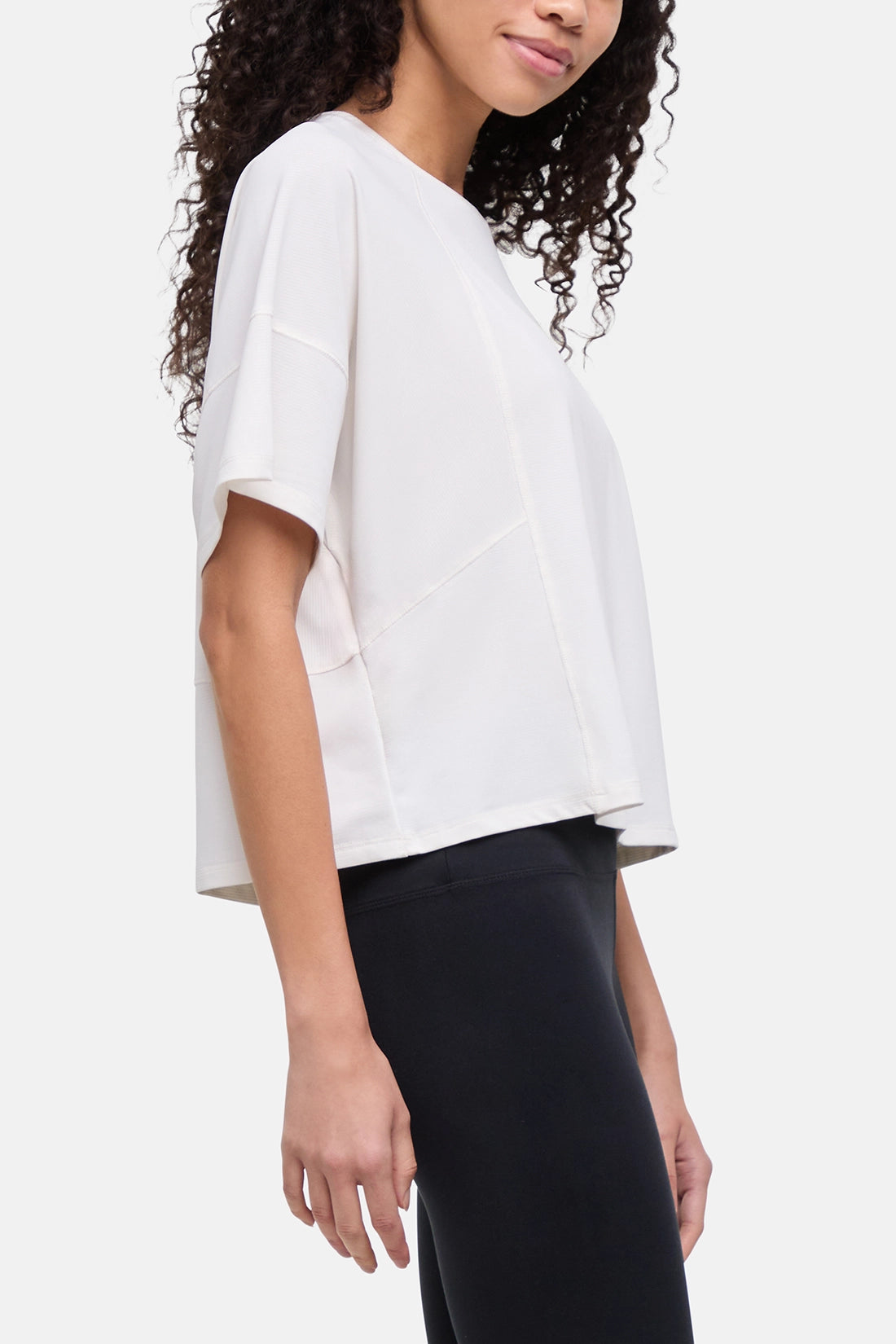 A woman wearing a white short-sleeve crop top with a relaxed, oversized fit and black leggings, shown from the side against a light gray studio background.  