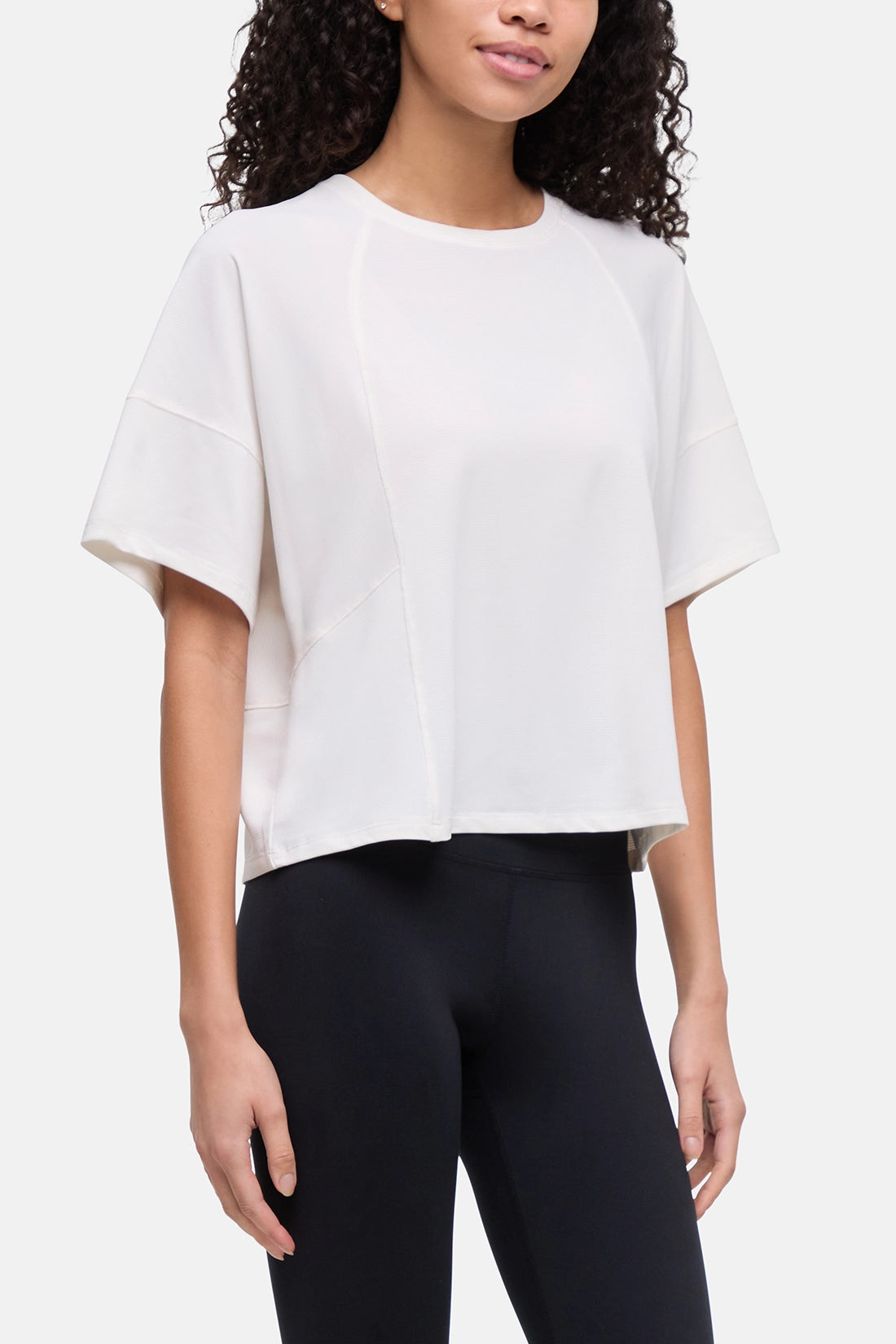 A woman wearing a white short-sleeve crop top with unique seam detailing and black leggings, shown from the waist up against a light gray studio background.  