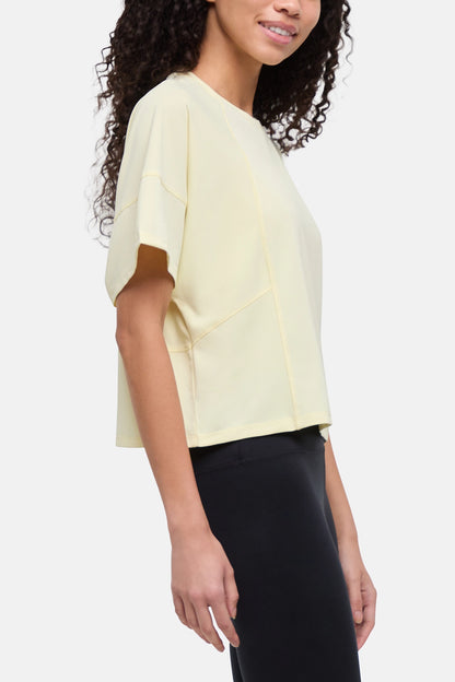 A woman wearing a pale yellow short-sleeve crop top with brown contrast panels on the sleeves, paired with black pants, shown against a light gray studio background. 