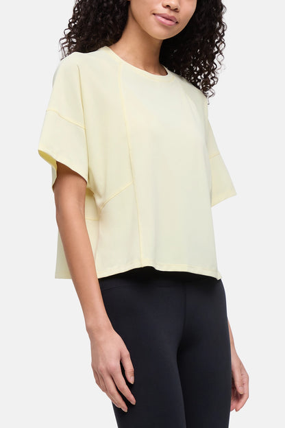 A woman wearing a pale yellow short-sleeve top with draped, layered sleeves and black leggings, photographed against a clean light gray studio background. 