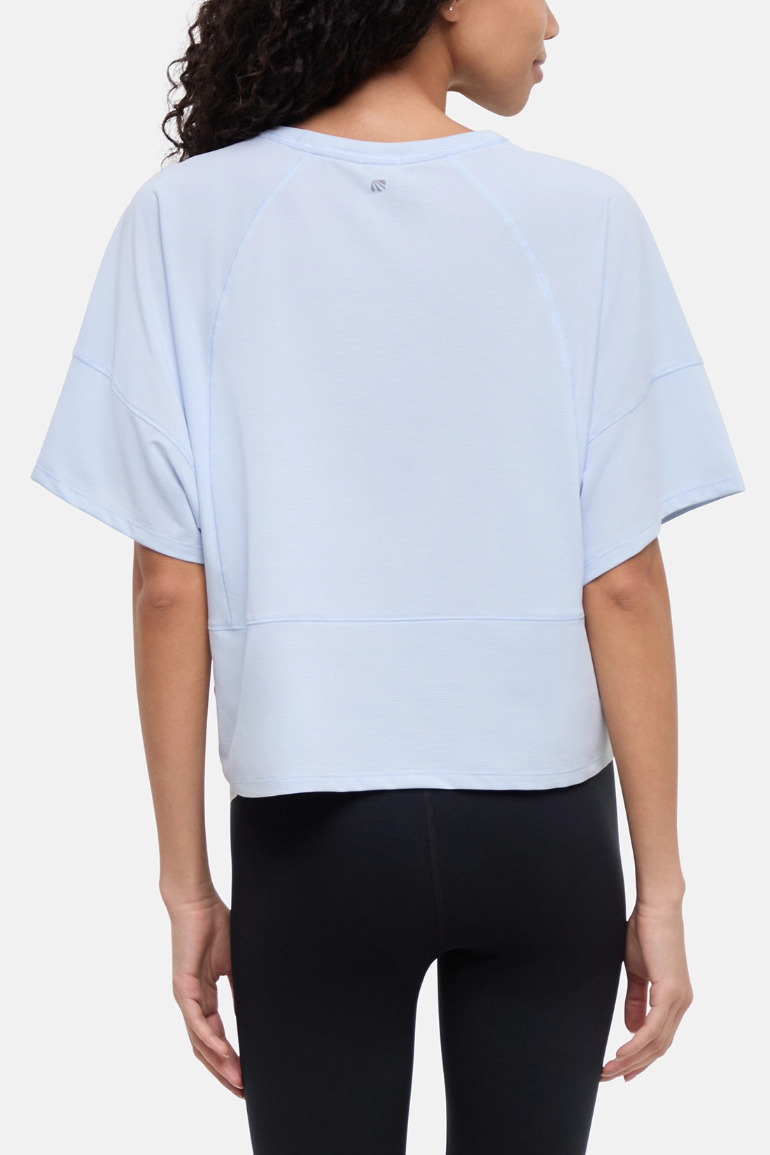 A woman shown from the back wearing a light blue cropped athletic top with raglan sleeves and black leggings, against a plain light gray studio background. 