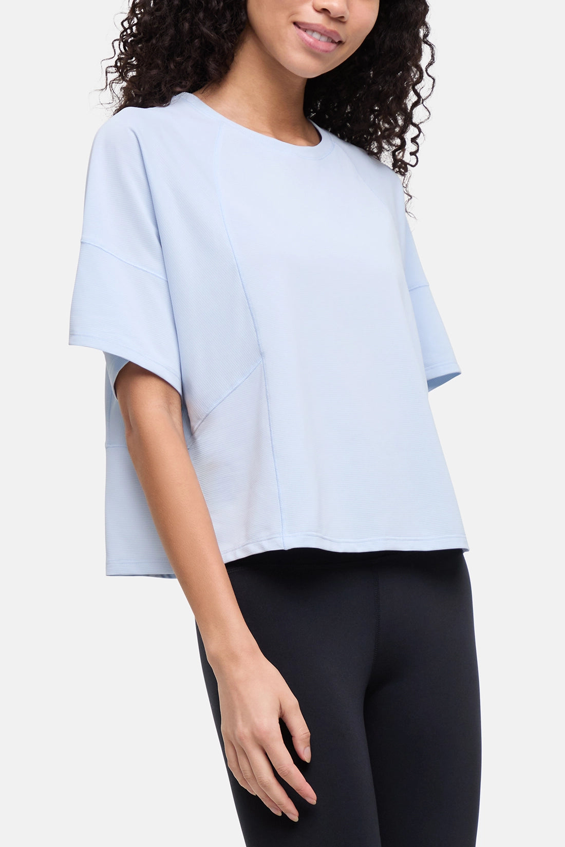 A woman wearing a light blue short-sleeve crop top with a relaxed, oversized fit and black leggings, shown against a plain light gray studio background. 