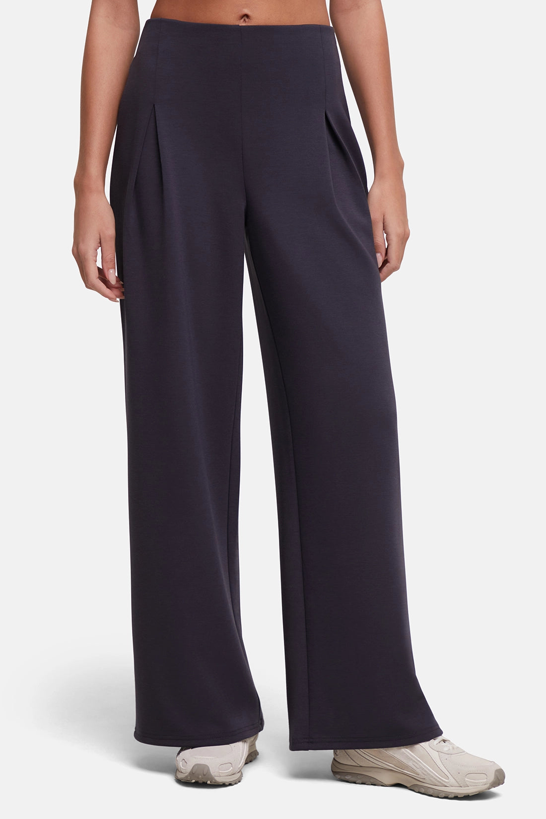 A model wearing dark navy wide-leg pleated trousers and light beige sneakers, shown from the waist down against a light gray studio background. 