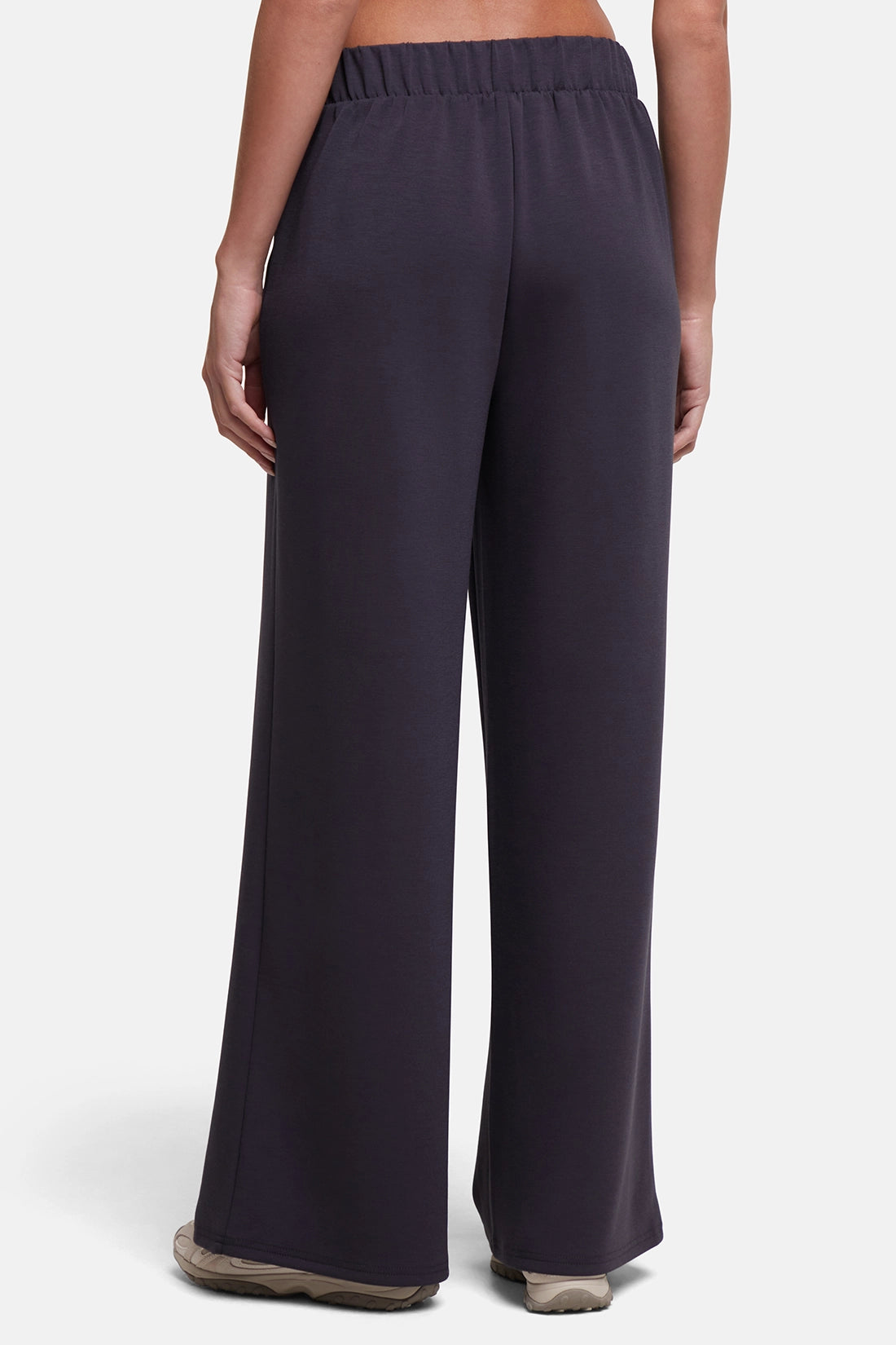 A person shown from the back wearing dark charcoal wide-leg pants with an elastic waistband, standing against a light gray studio background. 