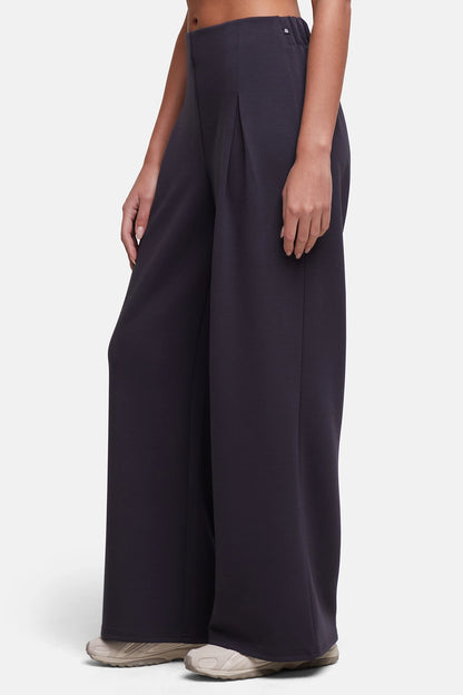 A model wearing dark charcoal wide-leg pleated trousers and light beige sneakers, shown from the waist down against a light gray studio background. 