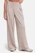 A model wearing high-waisted, wide-leg pleated trousers in a light taupe-beige color with front pleat detail, paired with light-colored sneakers, against a plain light gray studio background. 