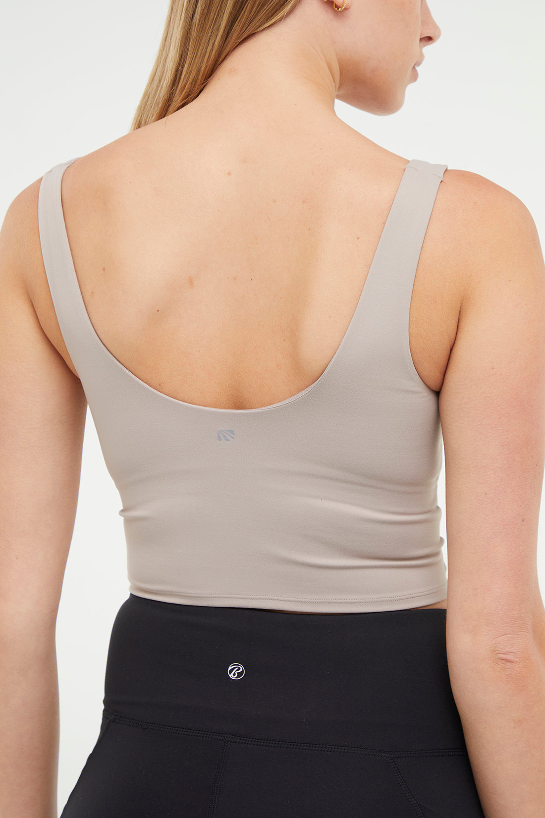 Close up back view of woman wearing a beige sports bra and black leggings on a white background. 