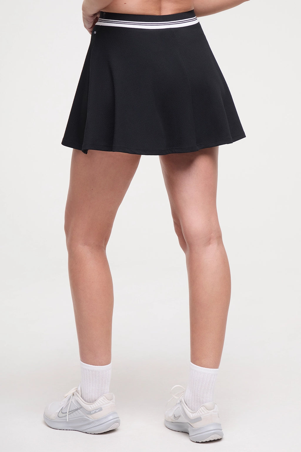 Back view of black tennis skirt with white stripe waistband worn by a woman on a white background. #color_black