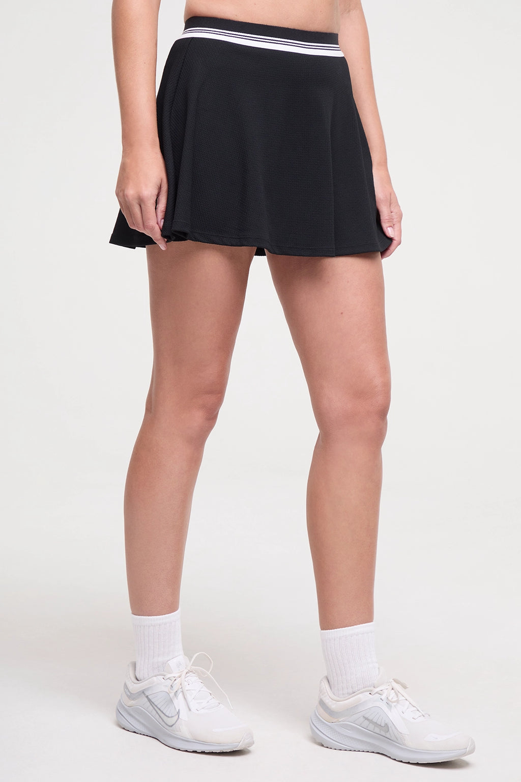 Side view of black tennis skirt with white stripe waistband worn by a woman on a white background. #color_black