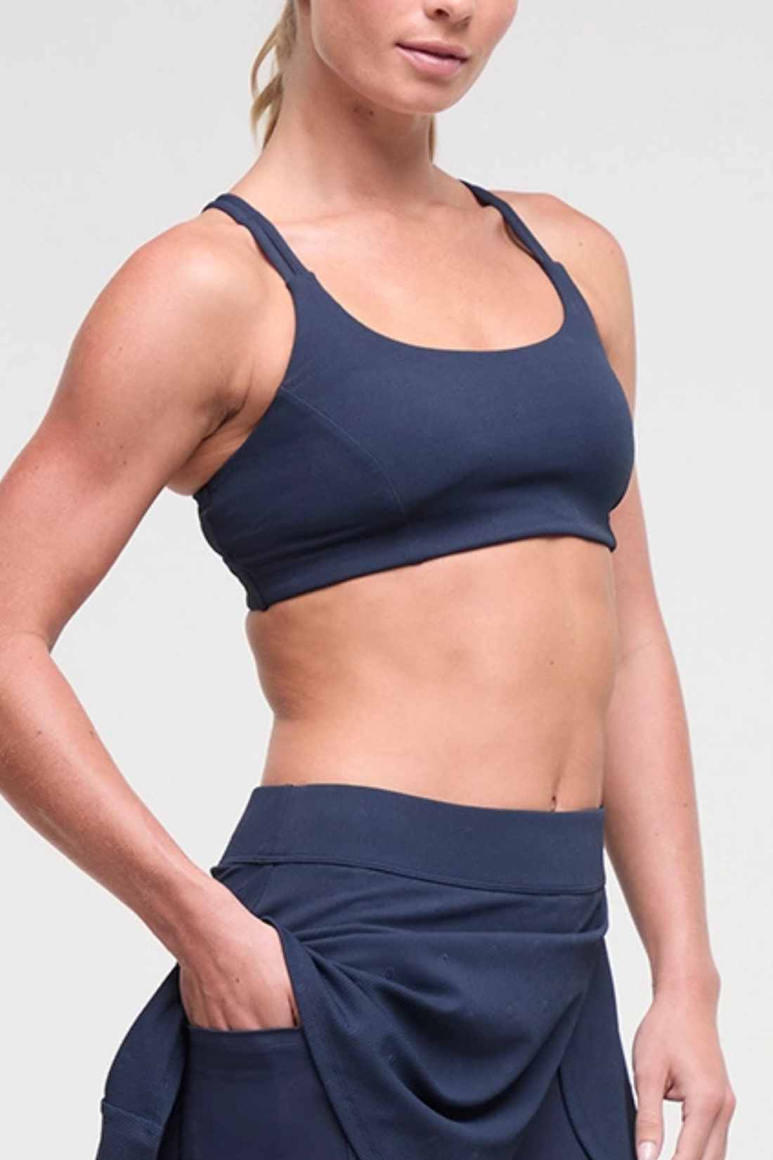 Side view of woman wearing a navy blue sports bra and skirt on a light gray background. 