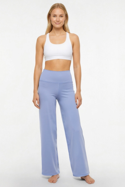 A woman modeling a two-piece activewear set consisting of a white sports bra and light periwinkle blue high-waisted wide-leg yoga pants, standing against a plain white studio background. 