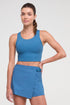 A woman modeling a matching athletic set consisting of a blue scoop-neck crop top and a blue wrap-style mini skirt, photographed against a light gray studio background. 