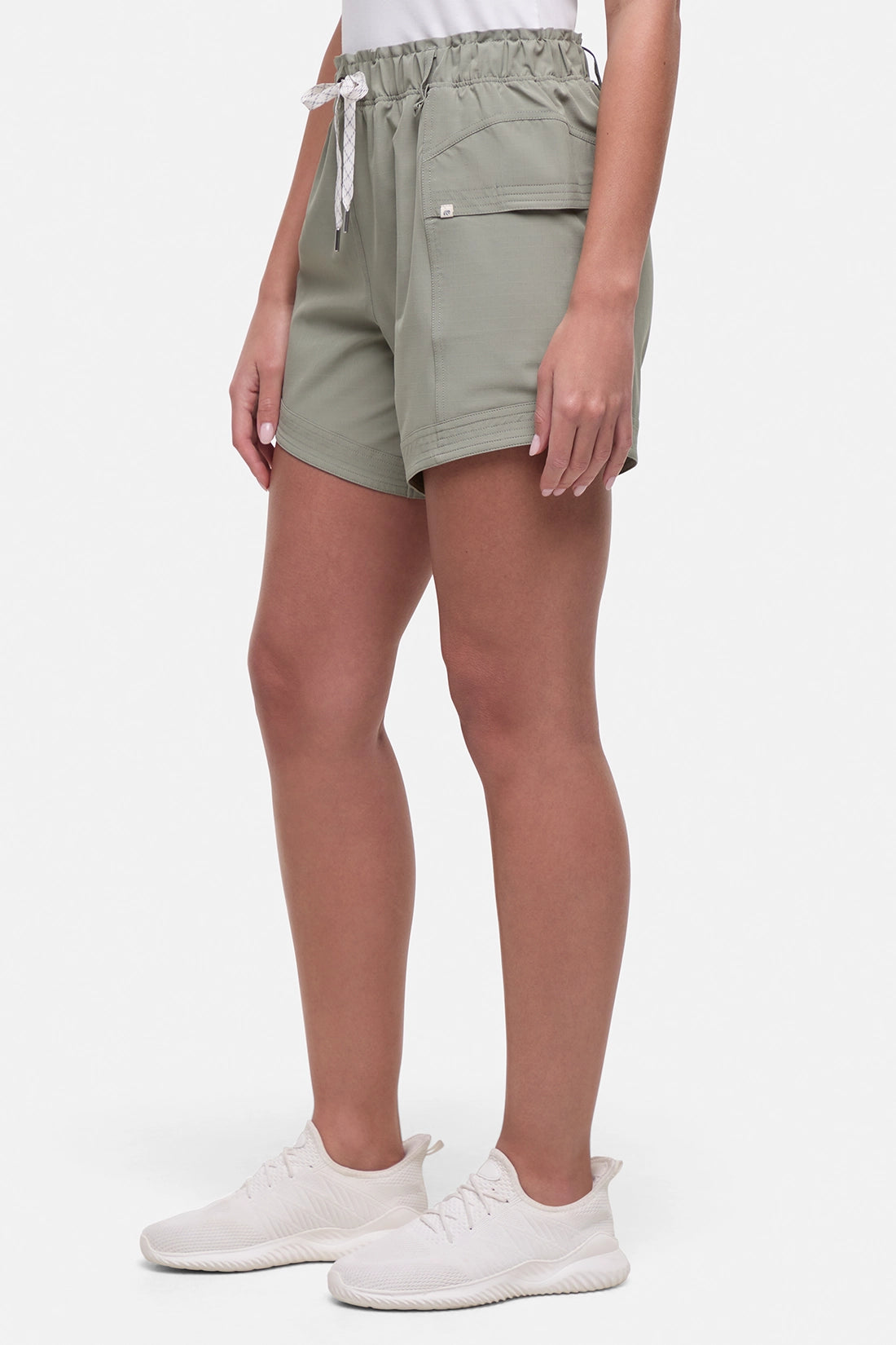 Side view of woman wearing sage green shorts with white drawstring waist and white sneakers on a white background. 